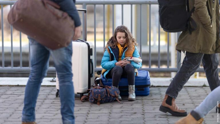 Sad Ukrainian immigrant child with luggage waiting at train station, Ukrainian war concept.